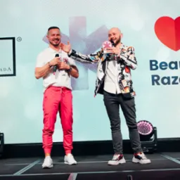 Dwaj mężczyźni na scenie, jeden w r&oacute;żowych spodniach i białem koszulce, drugi w czarno-białej marynarce w graficzne wzory, obaj trzymają mikrofony i uśmiechają się na tle ekranu z logo i napisem...