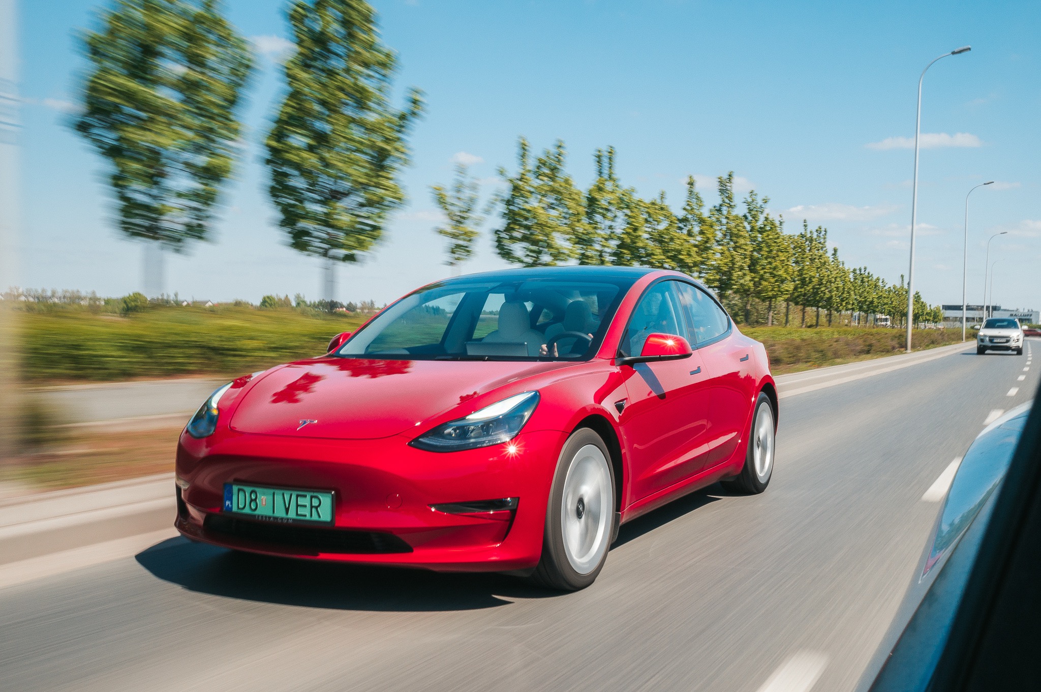 Czerwona Tesla Model 3 w ruchu na drodze, z zielonymi drzewami i niebieskim niebem w tle, uchwycona w dynamicznym ujęciu.