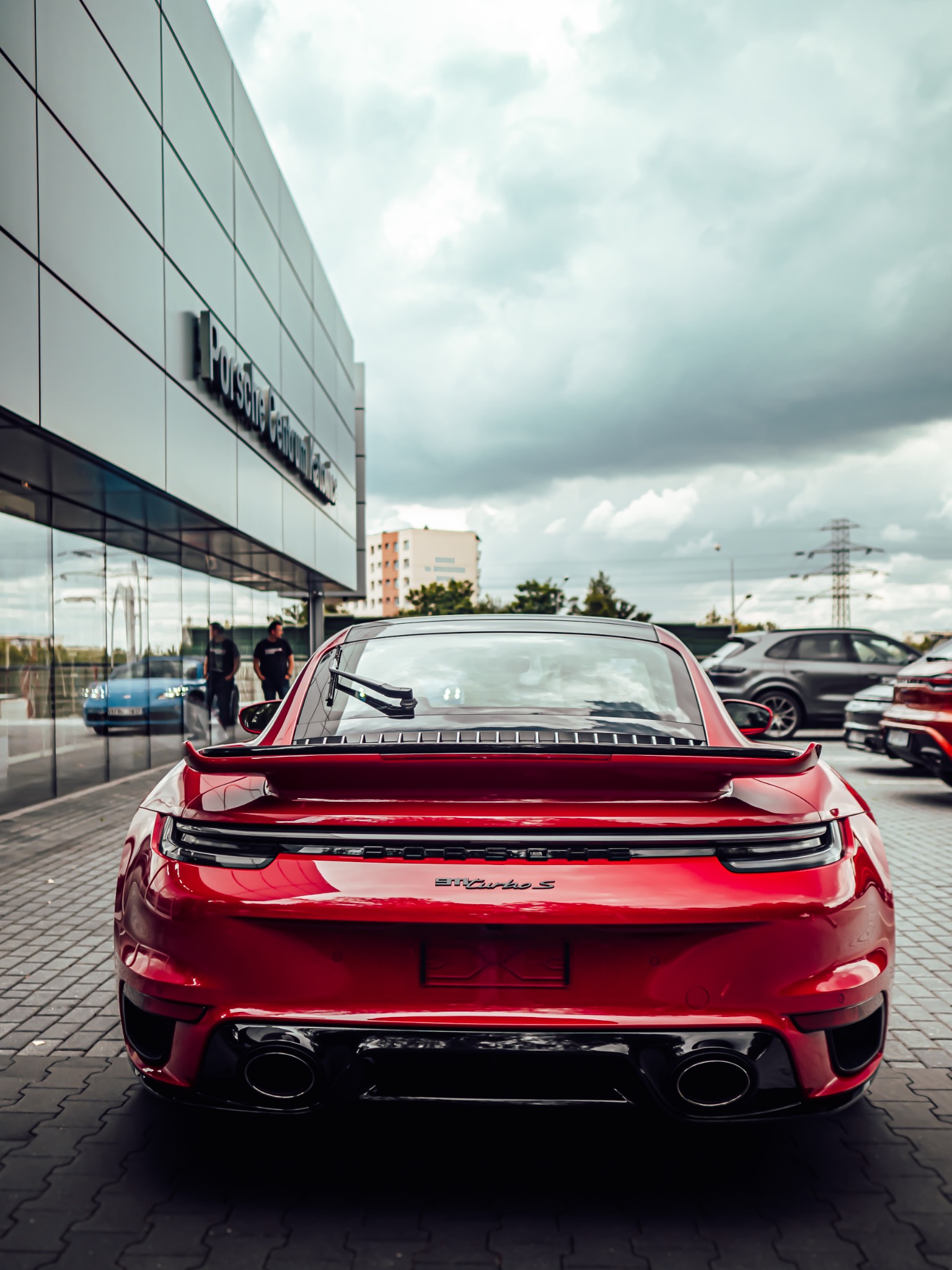 Czerwone Porsche 911 Turbo S zaparkowane przed salonem Porsche Centrum, widok z tyłu, zarys sylwetek w odbiciu szyby.