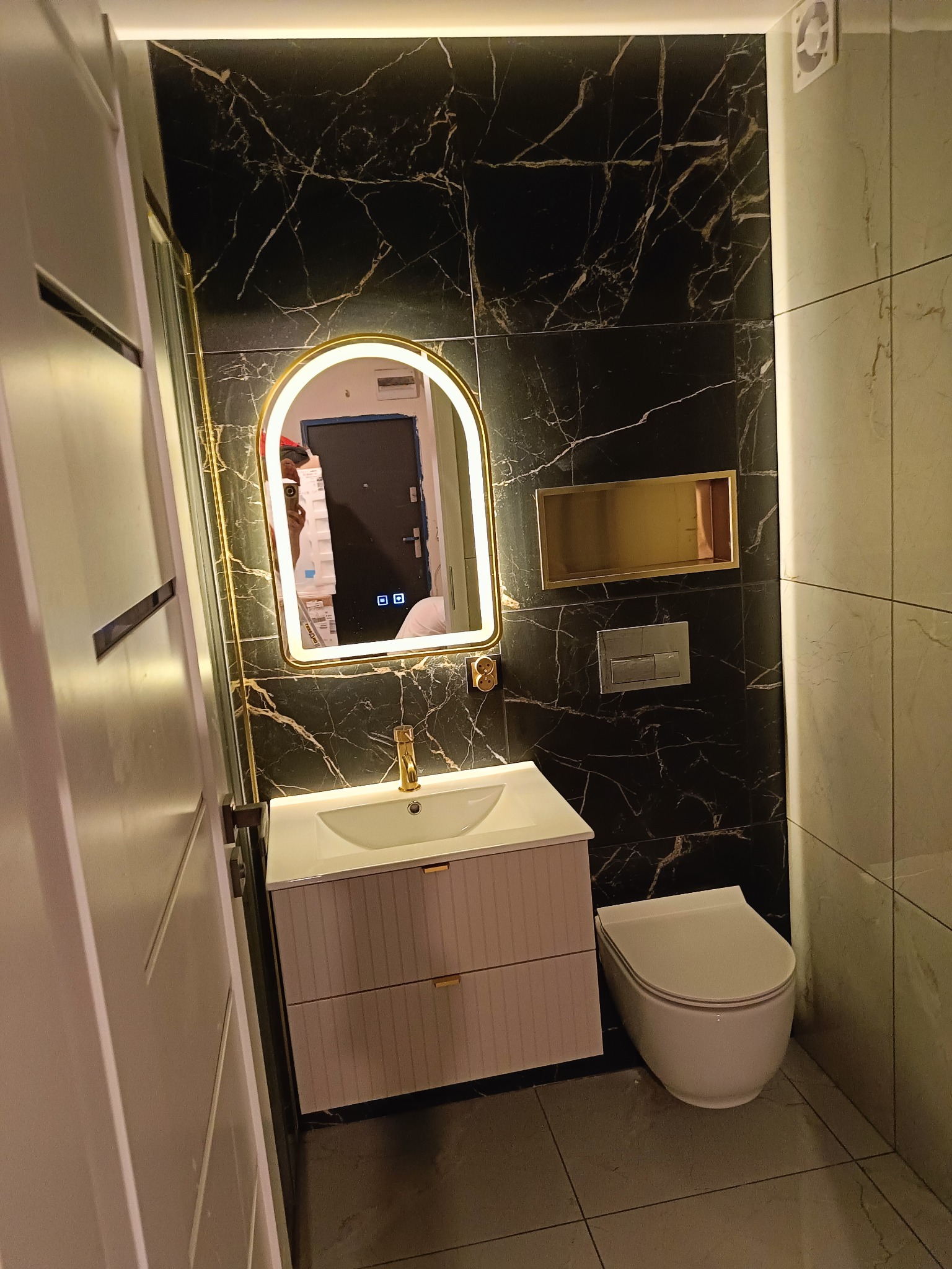Nowoczesna łazienka z czarnymi płytkami imitującymi marmur, podświetlane lustro w złotej ramie, biała szafka pod umywalką z złotymi uchwytami i wisząca toaleta.