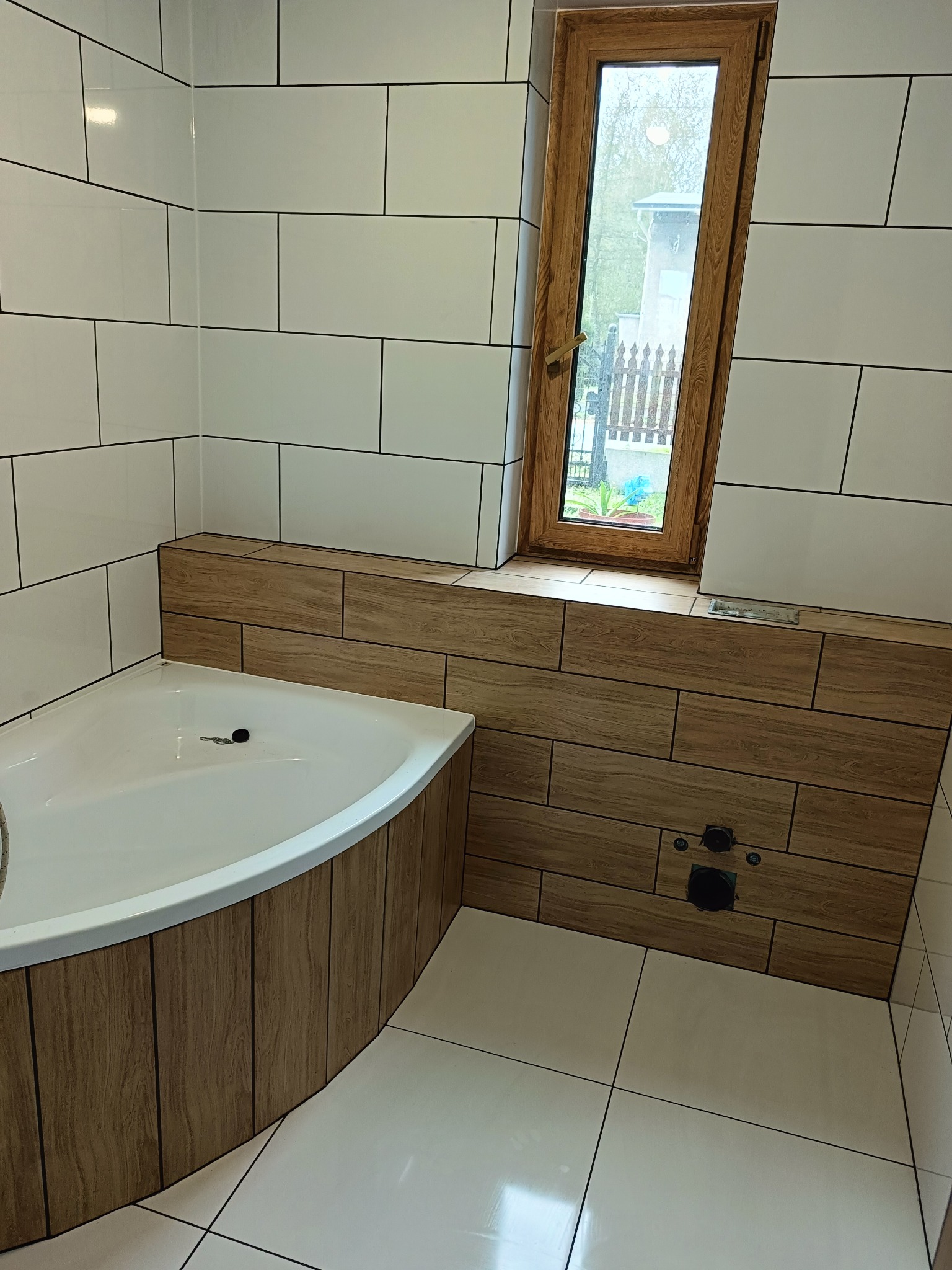 Nowoczesna łazienka po remoncie: białe płytki ścienne z czarną fugą, drewnopodobna obudowa wanny i okna, biała narożna wanna, widok na zewnątrz przez okno.