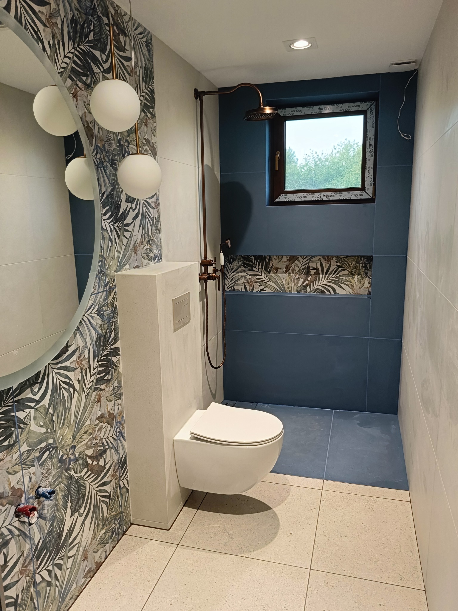 Wykończona łazienka w Knurowie z toaletą podwieszaną, kabiną prysznicową z deszczownicą w kolorze miedzi oraz okrągłym lustrem i płytkami w motywy roślinne.