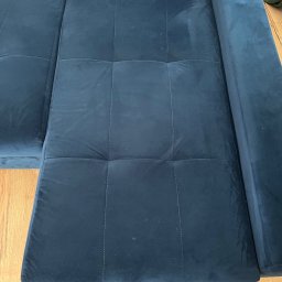 Mateusz Żylski - Fragment rozłożonej, ciemnoniebieskiej, welurowej sofy z widocznymi śladami czyszczenia na powierzchni, kwadratowe przeszycia, drewniana podłoga w tle.