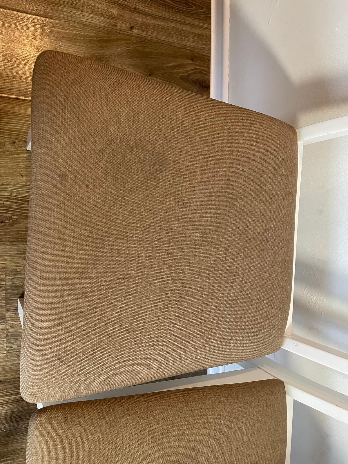 Widok z góry na krzesło z tapicerką w odcieniach beżu i białym stelażem, ustawione na podłodze imitującej drewno, z fragmentem białej ściany w tle.