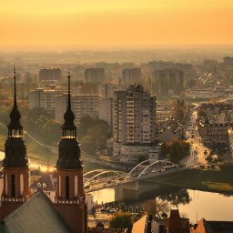 Adrian Mordarski - Panorama miasta o wschodzie słońca, z widokiem na rzekę, most i charakterystyczne wieże katedry, spowita delikatną mgłą.