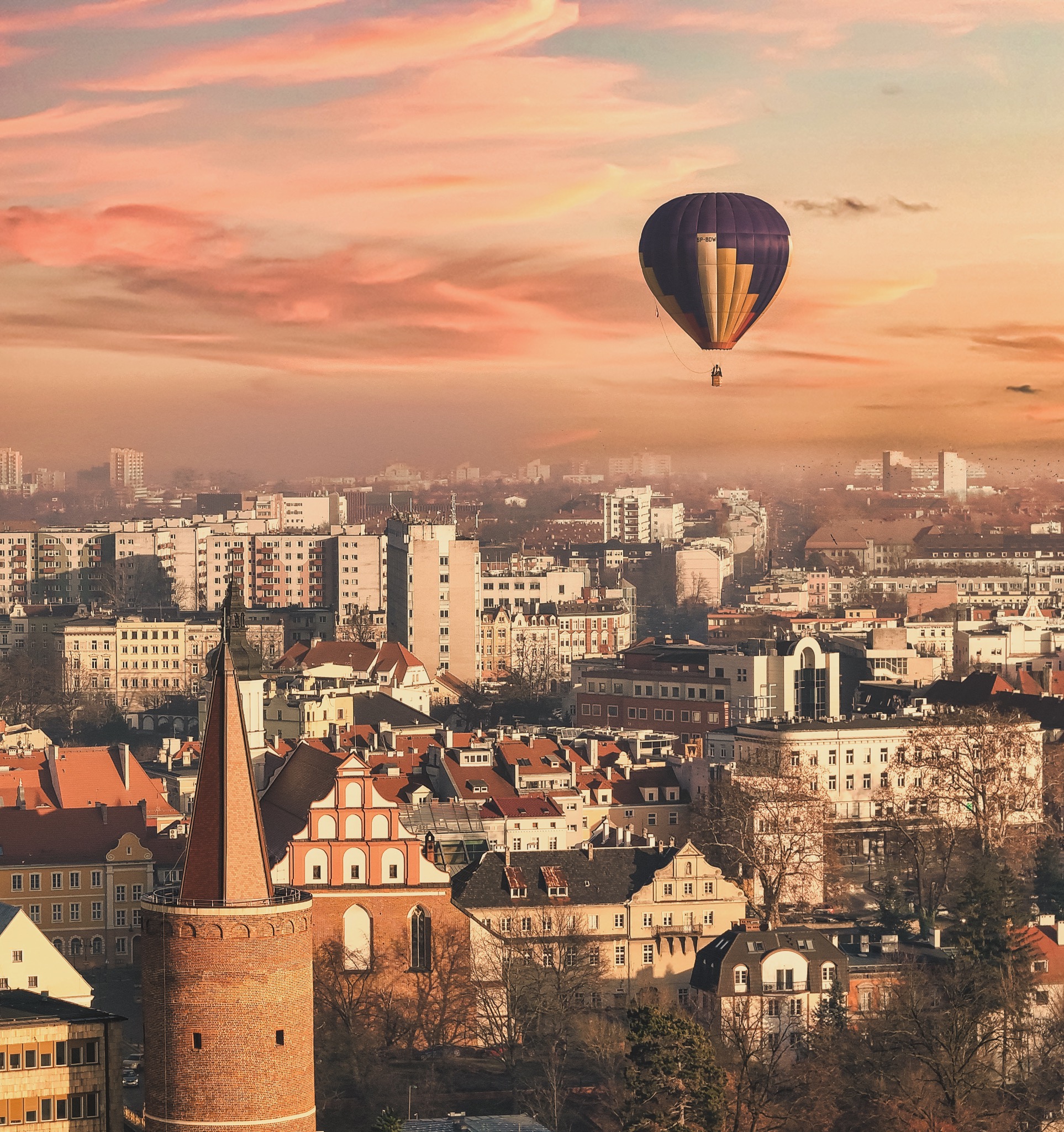Panorama miasta z ceglaną wieżą na pierwszym planie i balonem na tle różowego nieba o zachodzie słońca.