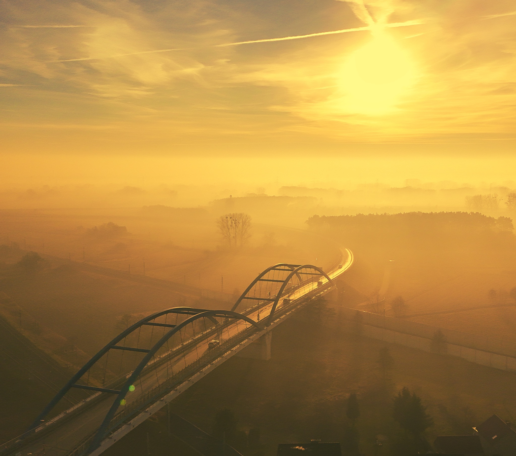 Poranny widok z lotu ptaka na most w Kępie spowity gęstą mgłą, oświetlony wschodzącym słońcem, tworzącym złocistą aurę.