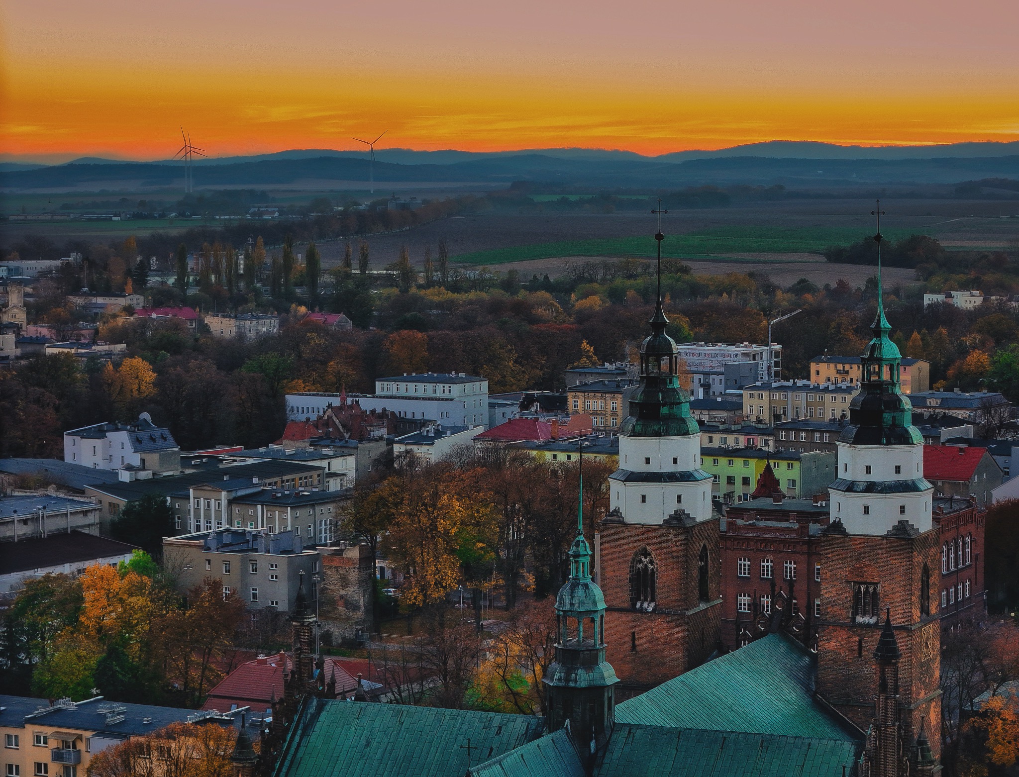 Panorama miasta z ceglanymi wieżami kościoła z zielonymi hełmami na pierwszym planie, w tle wzgórza i turbiny wiatrowe o zachodzie słońca.