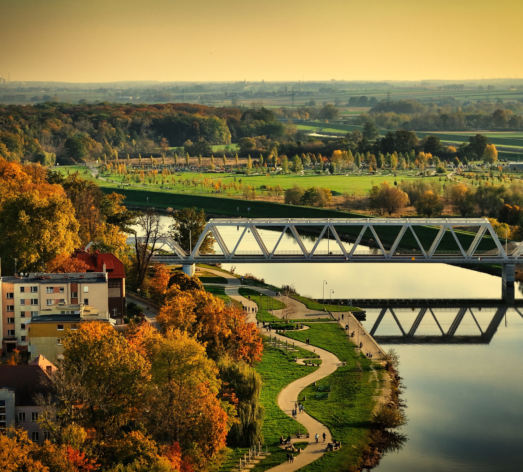 Jesienny krajobraz miejski z widokiem na rzekę, stalowy most kratownicowy, park z alejkami spacerowymi i budynki mieszkalne w ciepłym, popołudniowym świetle.