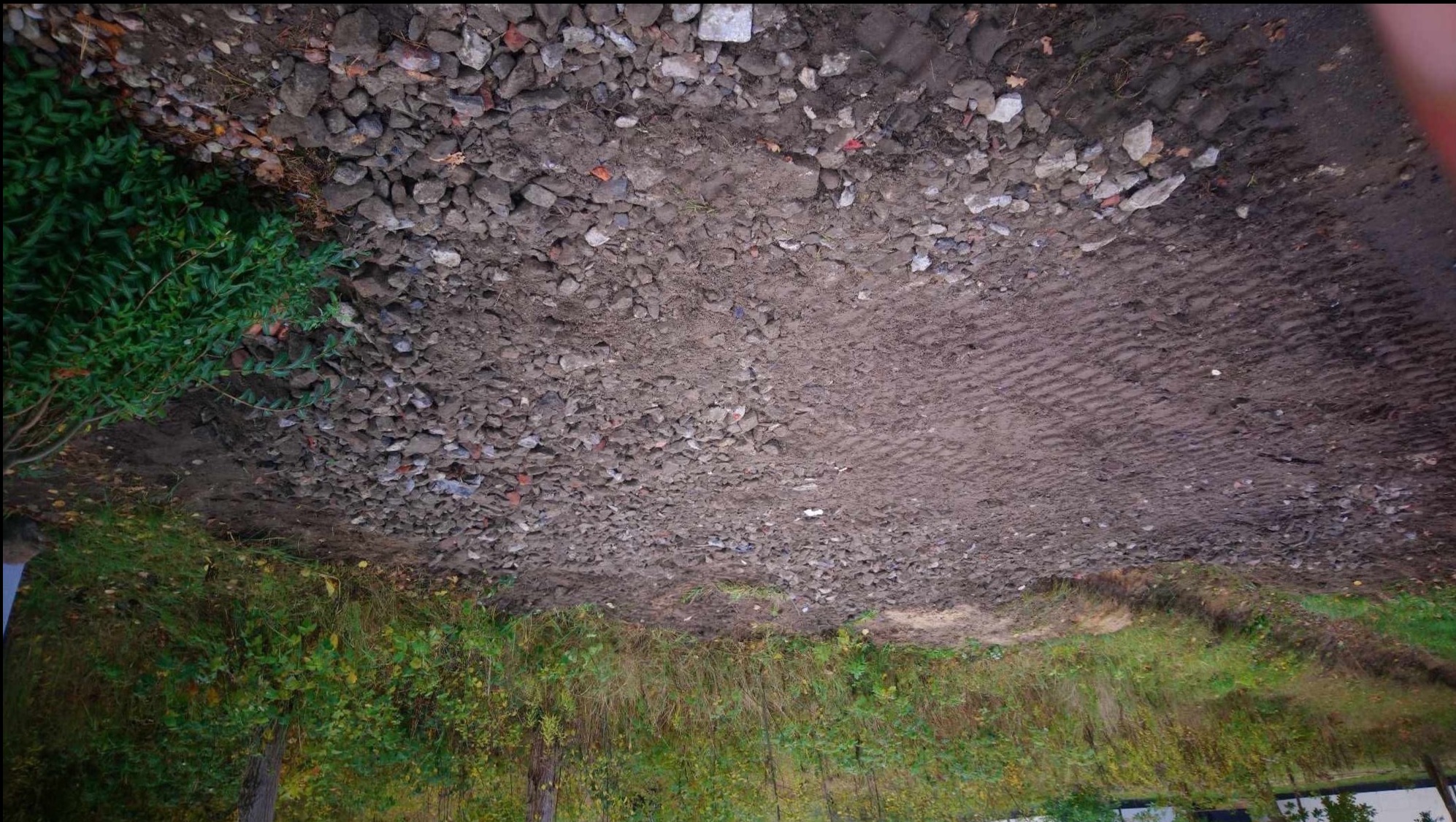 Ujęcie z góry na teren z ubitym podłożem i rozsypanym gruzem, widoczne ślady opon i roślinność na obrzeżach.