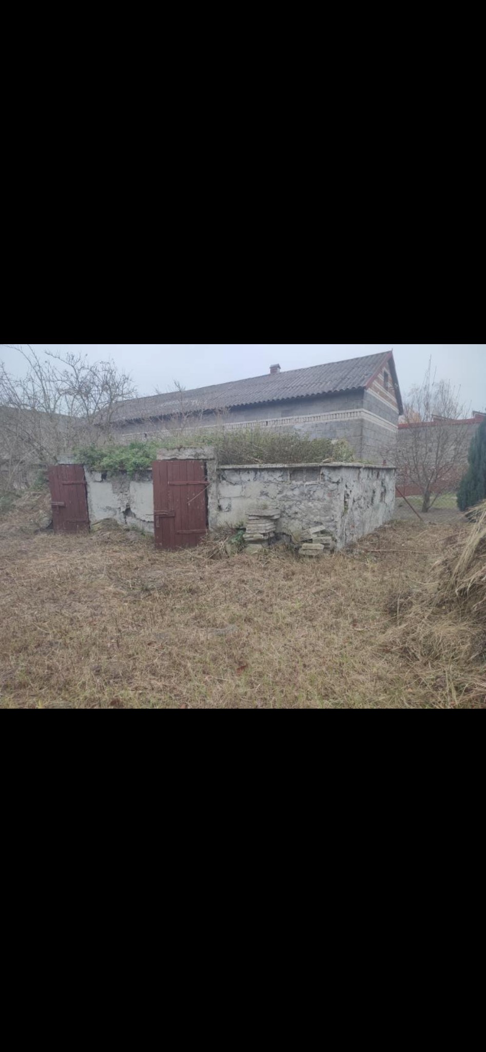 Stara, częściowo zburzona konstrukcja z kamienia i betonu z drewnianymi drzwiami na zarośniętym terenie, budynek w tle, pochmurny dzień.
