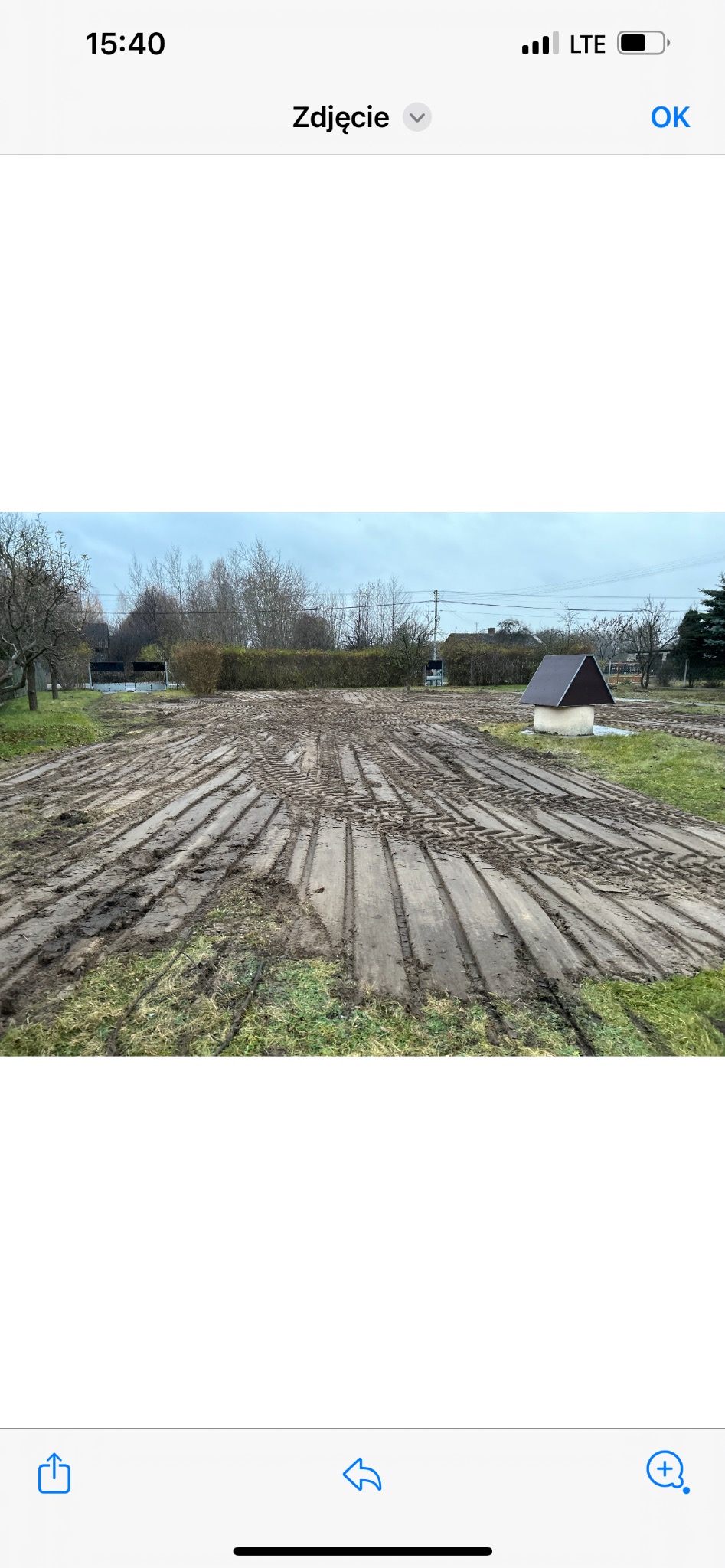 Przygotowany teren pod budowę z ułożonymi płytami drewnianymi, pokrytymi błotem, ślady opon, widoczna studnia z daszkiem w tle.