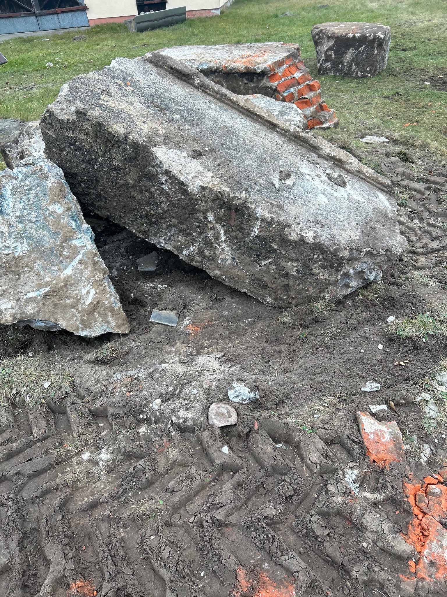 Sterta gruzu z betonu i cegieł na błotnistym terenie z wyraźnymi śladami opon ciężkiego sprzętu, pozostałości po rozbiórce, z fragmentami trawy i budynku w tle.