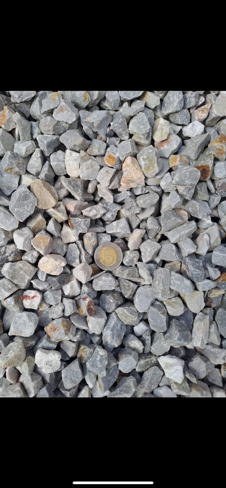 Szary tłuczeń o nieregularnych kształtach, z monetą 5 zł dla skali, widoczne drobne, brązowe przebarwienia na niektórych kamieniach.