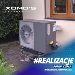 POLSKA EKOLOGIA SPÓŁKA Z OGRANICZONĄ ODPOWIEDZIALNOŚCIĄ - Szara pompa ciepła Weber zamontowana na metalowej podstawie przy ścianie domu, z widocznymi przewodami i fragmentem elewacji z oknem, otoczona zielenią.