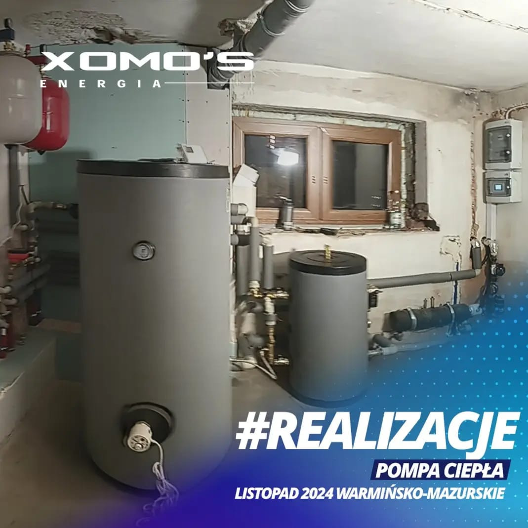 Szara pompa ciepła w piwnicy, z widocznymi rurami i oknem w tle, zainstalowana w listopadzie 2024 w województwie warmińsko-mazurskim, z logo firmy XOMO'S Energia.
