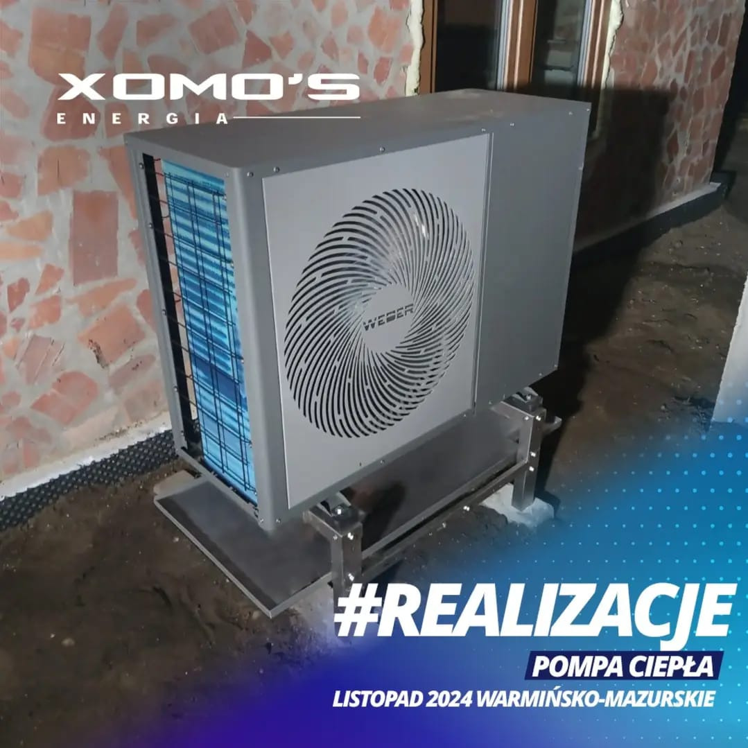 Szara pompa ciepła Weber na metalowej konstrukcji, zainstalowana przy ścianie budynku z cegły, z widocznym logo Xomo's Energia i napisem #REALIZACJE Listopad 2024 Warmińsko-Mazurskie.