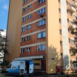 WASH WASH PIOTR REPLIN - Pracownik na linach myje elewację wysokiego budynku, na dole zaparkowany van z logo firmy świadczącej usługi czyszczenia, widoczny drugi pracownik obsługujący węże.