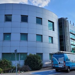 WASH WASH PIOTR REPLIN - Niebiesko-biały samochód dostawczy firmy sprzątającej, z logo 'Wash Wash', zaparkowany przed nowoczesnym, srebrnym budynkiem biurowym o zaokrąglonym kształcie i panelowej elewacji, w tle błękitne...