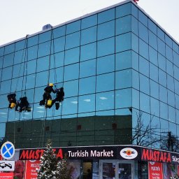 WASH WASH PIOTR REPLIN - Trzech alpinistów przemysłowych myje szklaną elewację budynku Mustafa Turkish Market w pochmurny, zimowy dzień. Widoczne drzewa i śnieg.