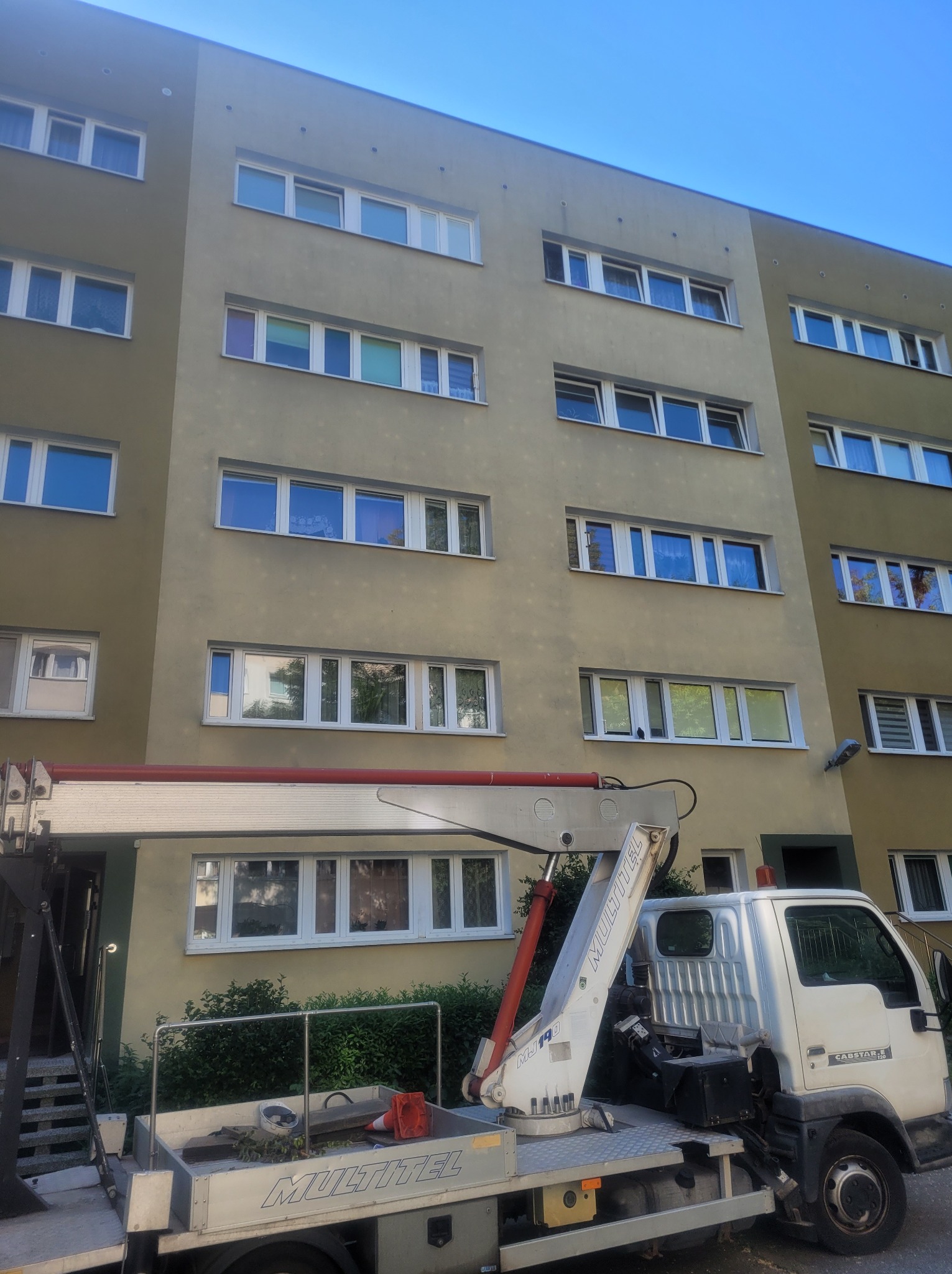 Ciężarówka z podnośnikiem hydraulicznym na tle bloku mieszkalnego z oknami; widoczny fragment elewacji i niebo