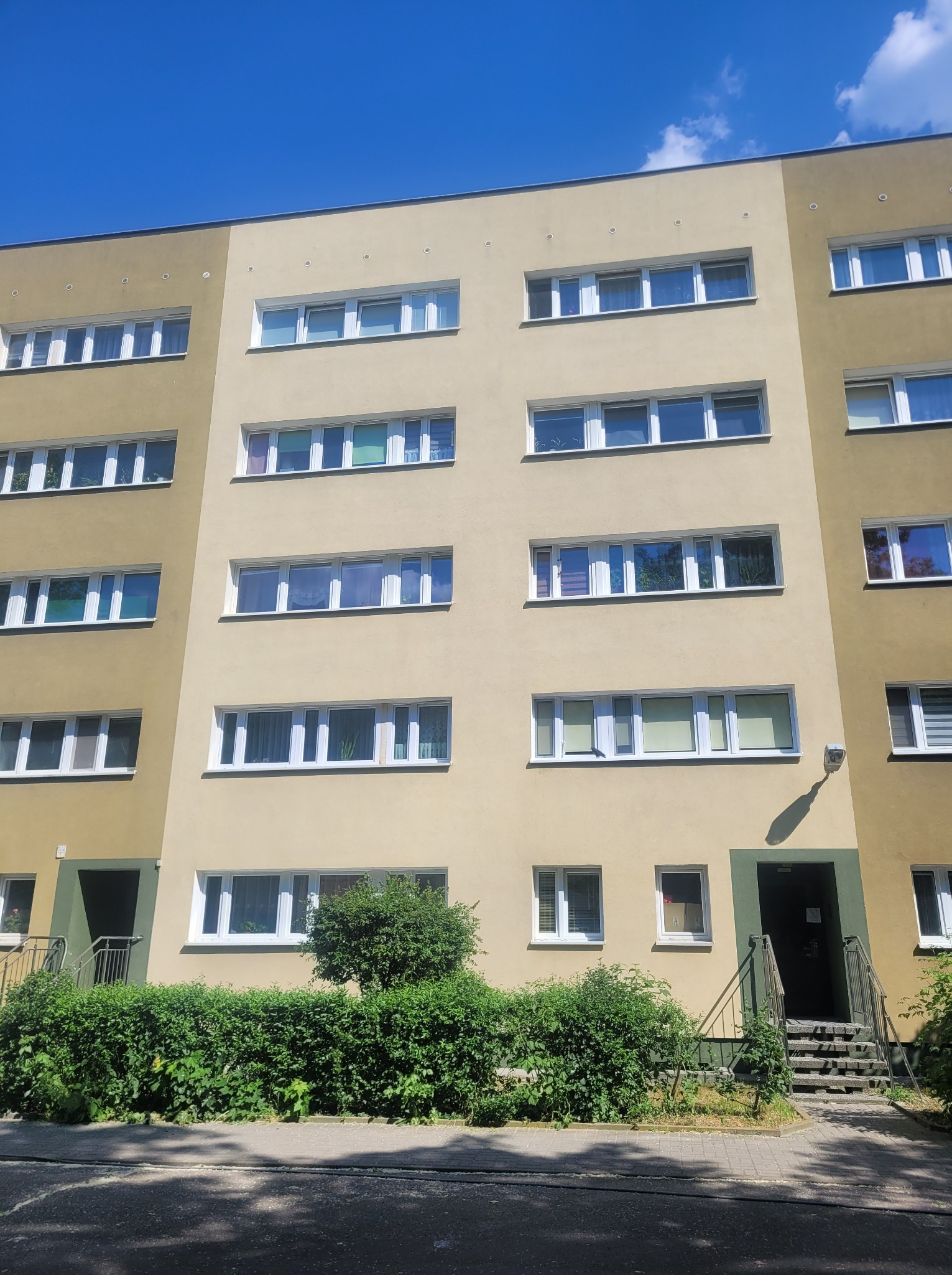 Wielopiętrowy budynek mieszkalny z elewacją w odcieniach beżu i oliwki, widoczny z poziomu ulicy w słoneczny dzień.