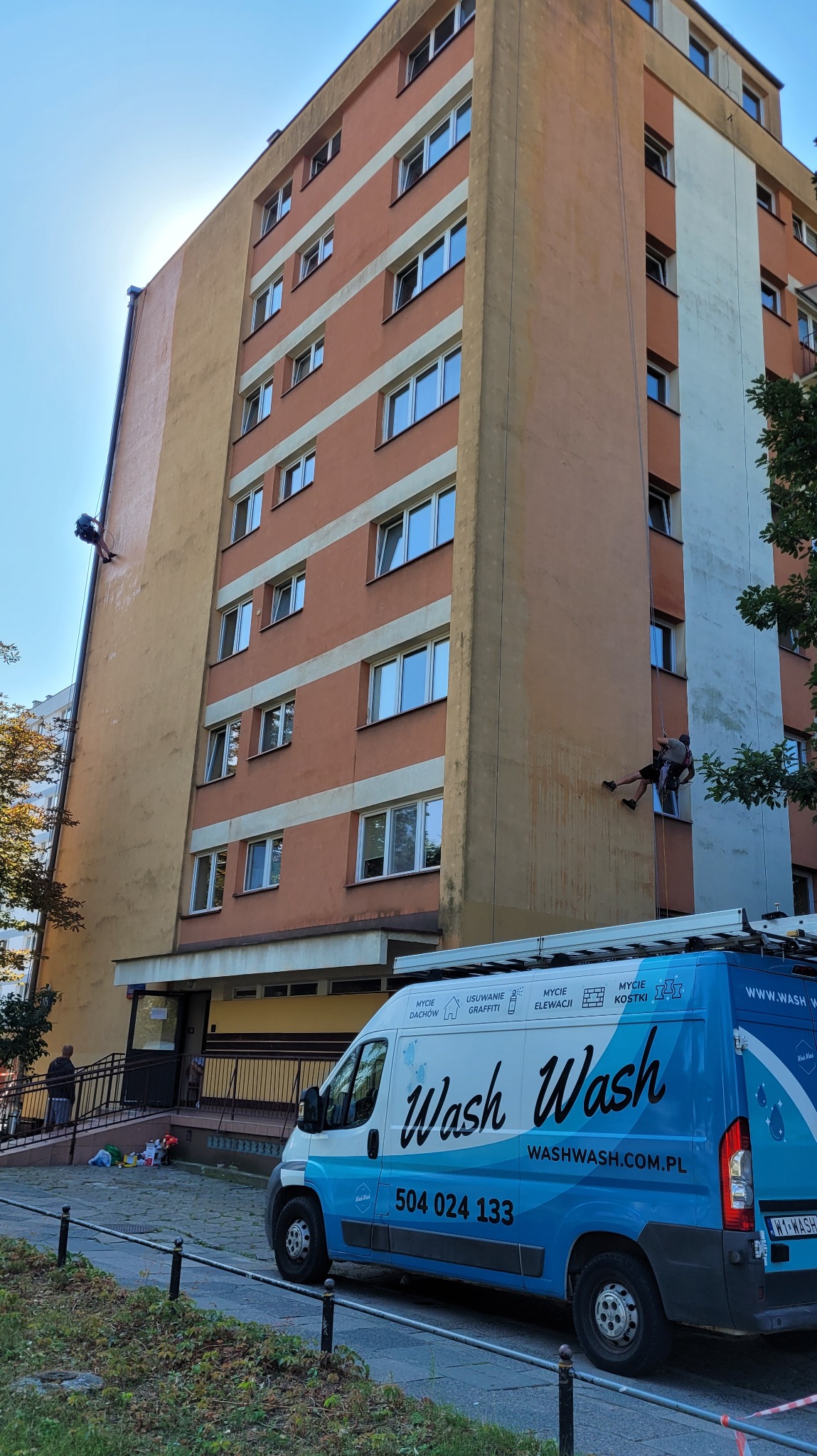 Pracownik na linach myje elewację wysokiego budynku, widoczny firmowy samochód z napisem 'Wash Wash' oraz numerem telefonu.