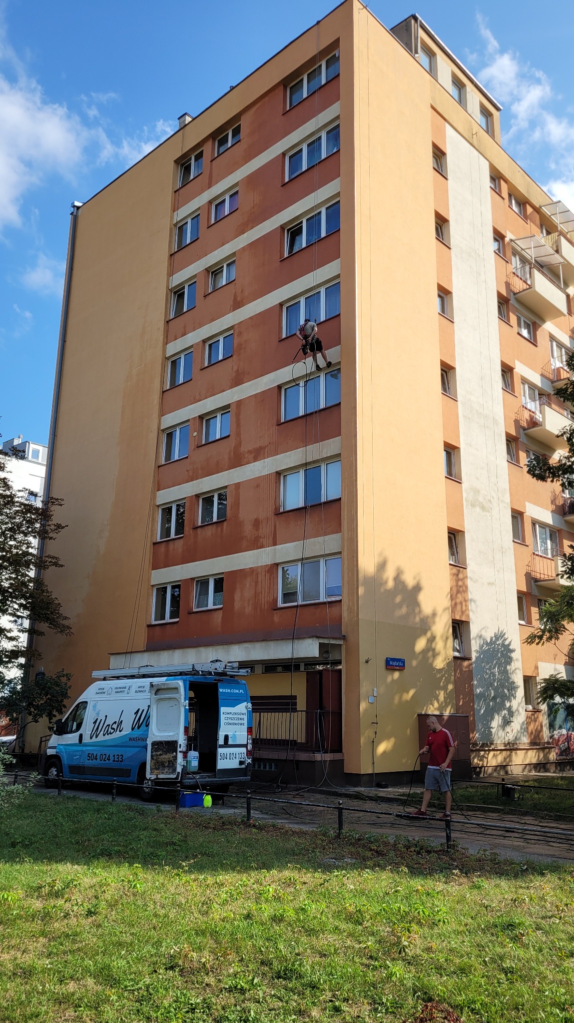 Pracownik na linach myje elewację wysokiego budynku, na dole zaparkowany van z logo firmy świadczącej usługi czyszczenia, widoczny drugi pracownik obsługujący węże.