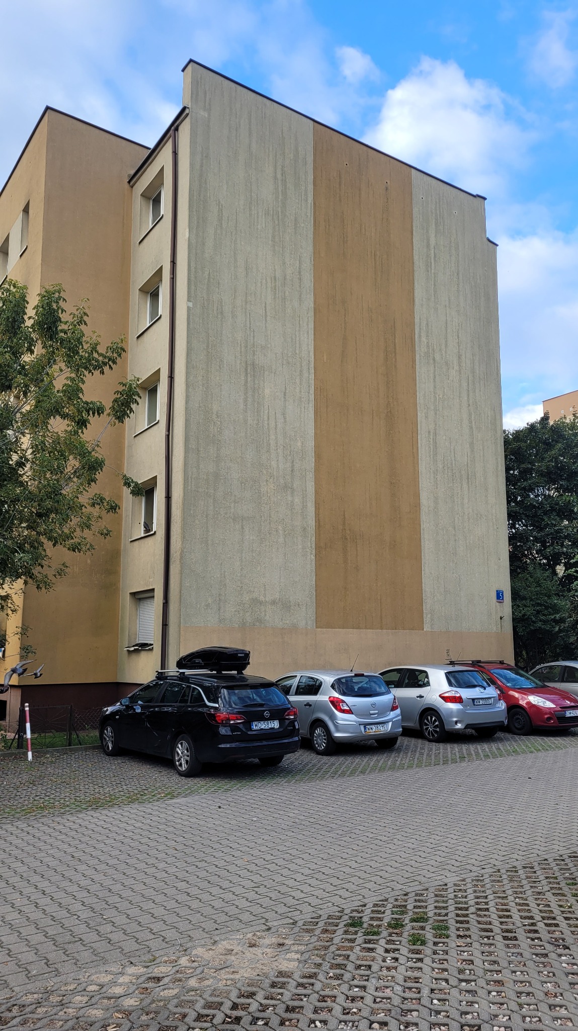 Wielopiętrowy budynek mieszkalny z elewacją w odcieniach beżu i szarości, widoczny z poziomu parkingu z kostki brukowej, gdzie zaparkowane są samochody.