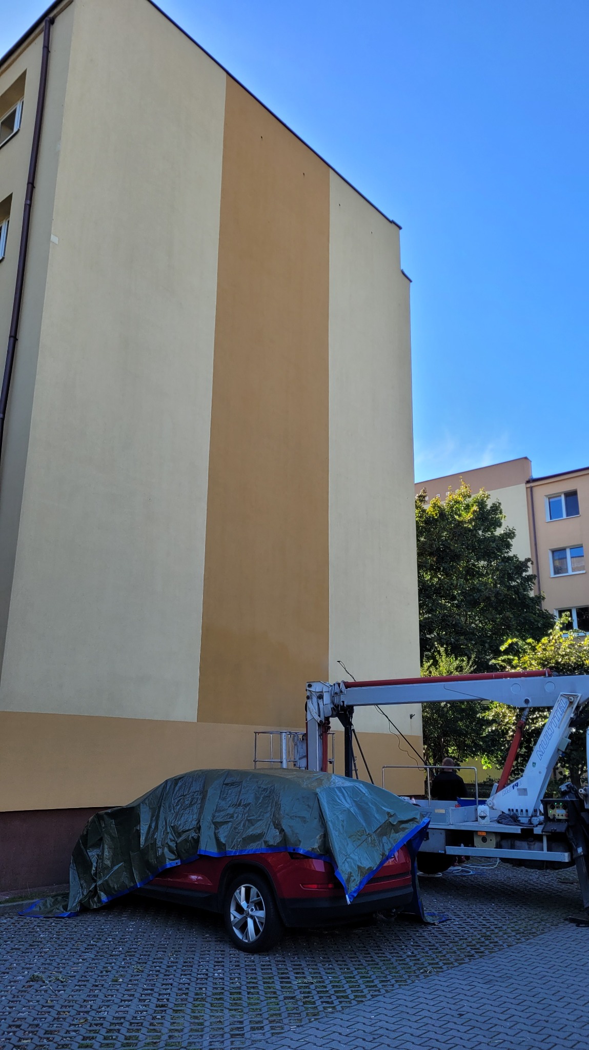 Wysoki budynek z częściowo pomalowaną elewacją w odcieniach beżu i brązu, zabezpieczony samochód pod plandeką i podnośnik koszowy obok, na kostce brukowej.