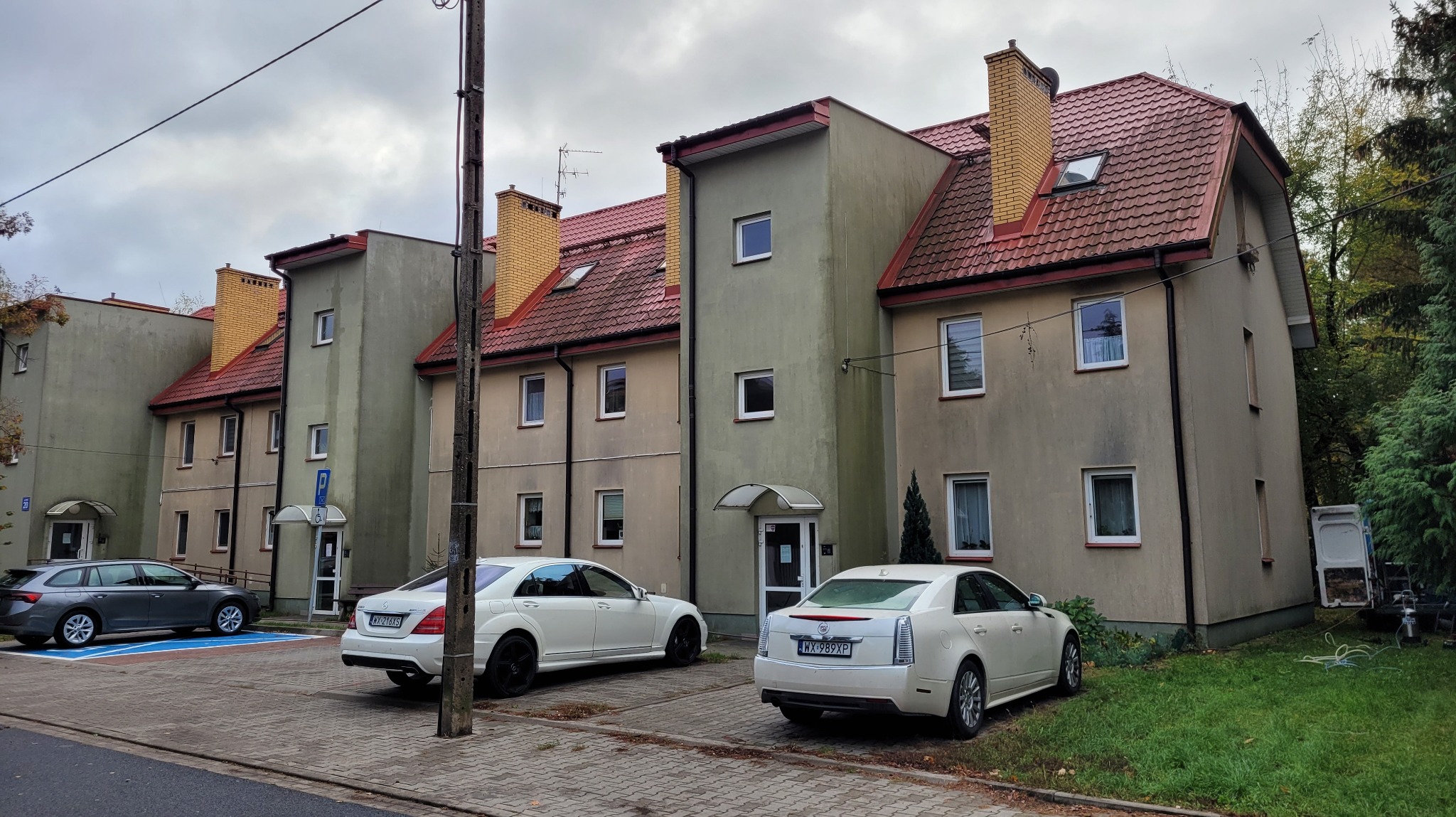 Trzysegmentowy budynek mieszkalny z czerwonym dachem, elewacją w odcieniach beżu i zieleni, widoczny z poziomu chodnika, z zaparkowanymi samochodami i słupem energetycznym.