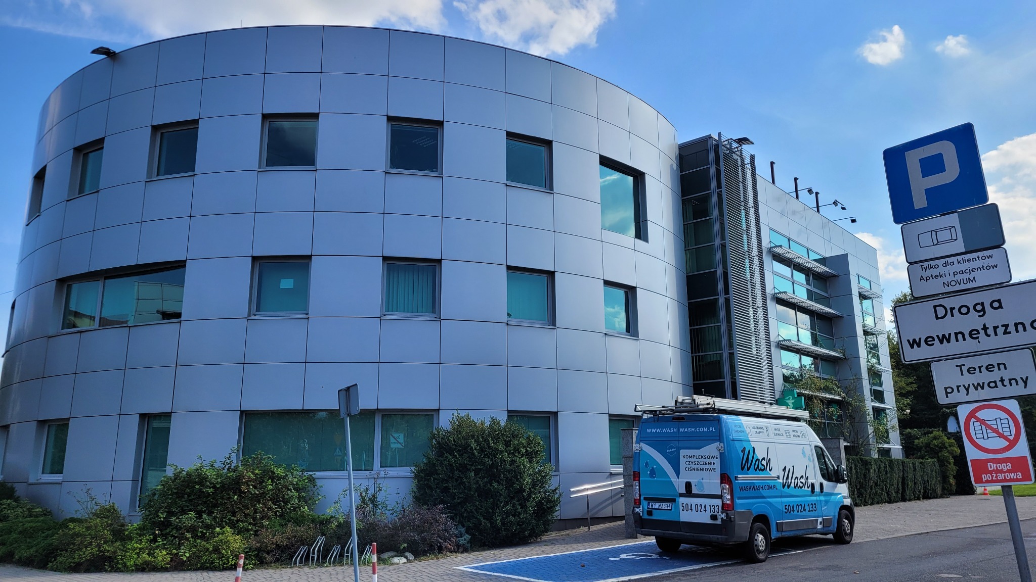 Niebiesko-biały samochód dostawczy firmy sprzątającej, z logo 'Wash Wash', zaparkowany przed nowoczesnym, srebrnym budynkiem biurowym o zaokrąglonym kształcie i panelowej elewacji, w tle błękitne...