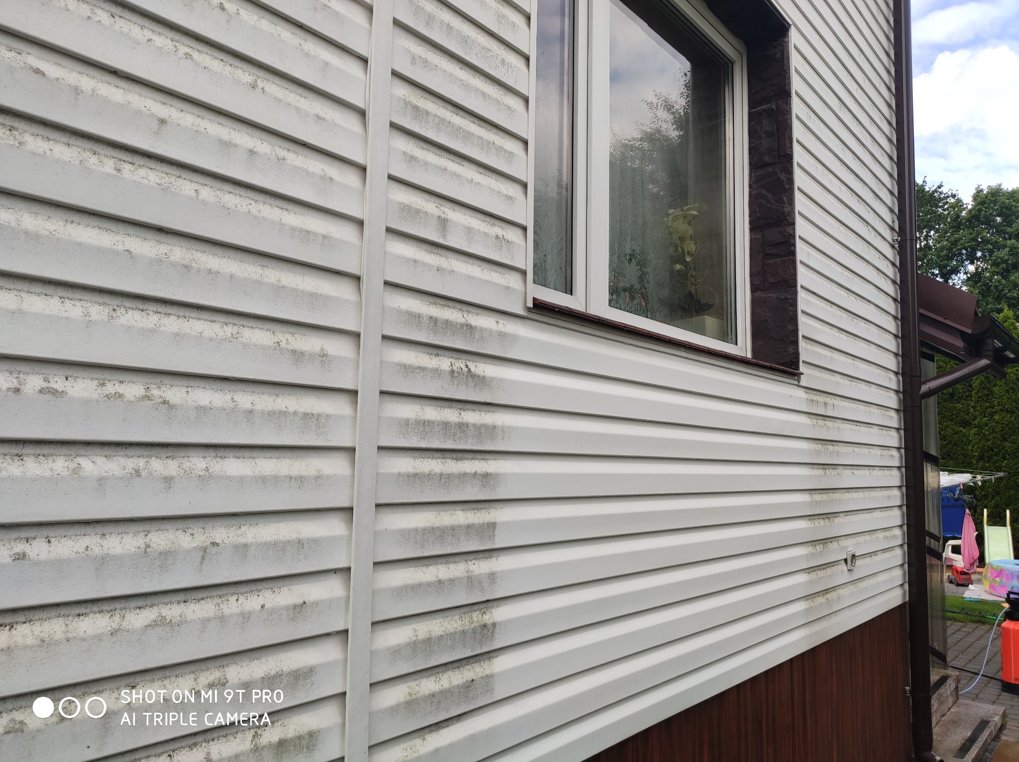 Fragment białej elewacji sidingowej z widoczną różnicą w czystości, po lewej stronie brudna, po prawej umyta, okno z brązową ramą i widoczną rośliną wewnątrz, na dole brązowa podmurówka.