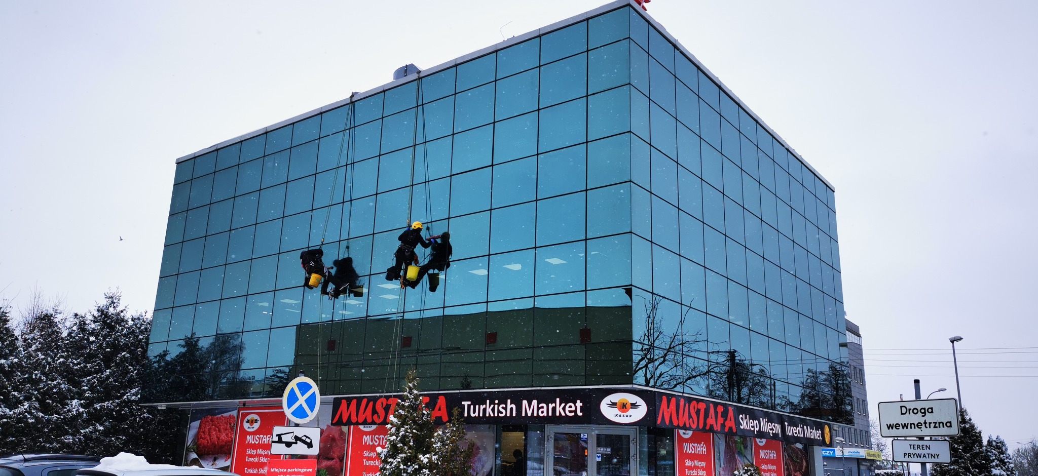 Trzech alpinistów przemysłowych myje szklaną elewację budynku Mustafa Turkish Market w pochmurny, zimowy dzień. Widoczne drzewa i śnieg.