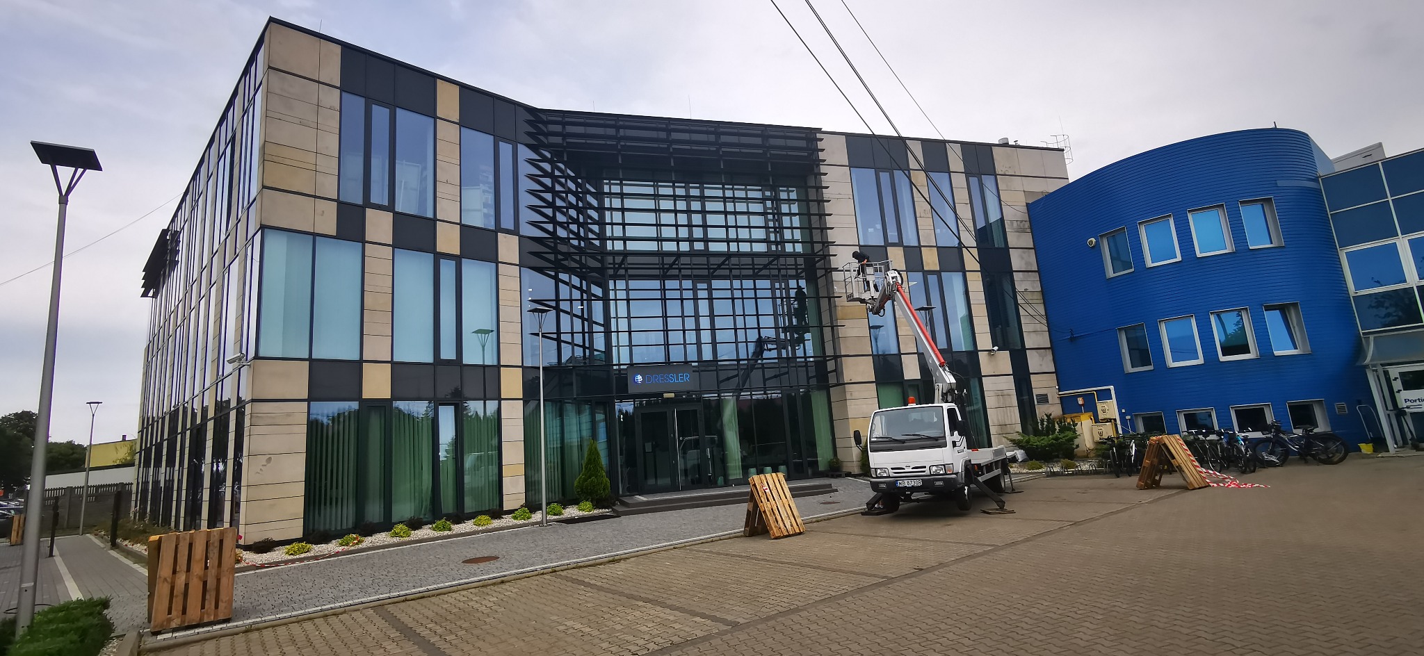 Pracownik na podnośniku koszowym czyści elewację biurowca z elementami szkła i kamienia, widoczny biały samochód dostawczy i zaparkowane rowery.