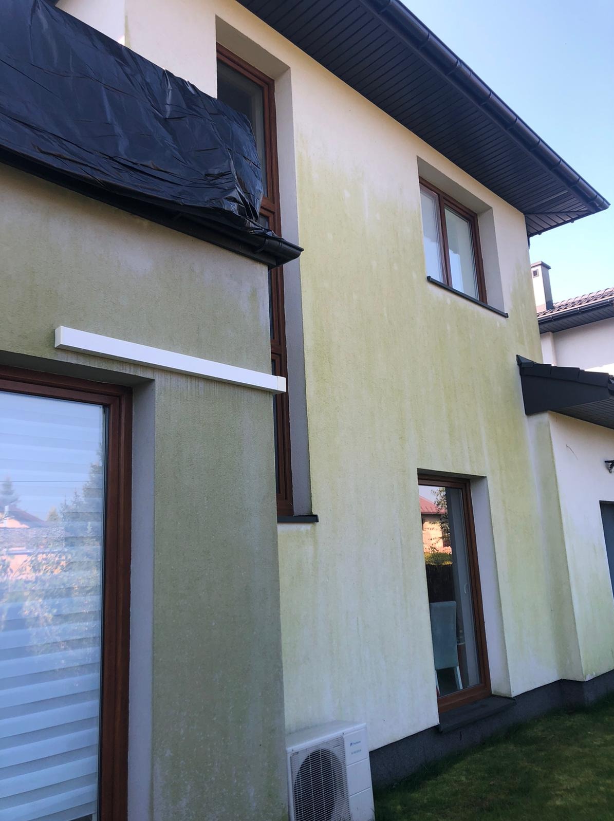 Elewacja dwukondygnacyjnego domu z widocznymi zielonymi zabrudzeniami, częściowo zakryta czarną folią ochronną, przygotowana do czyszczenia.