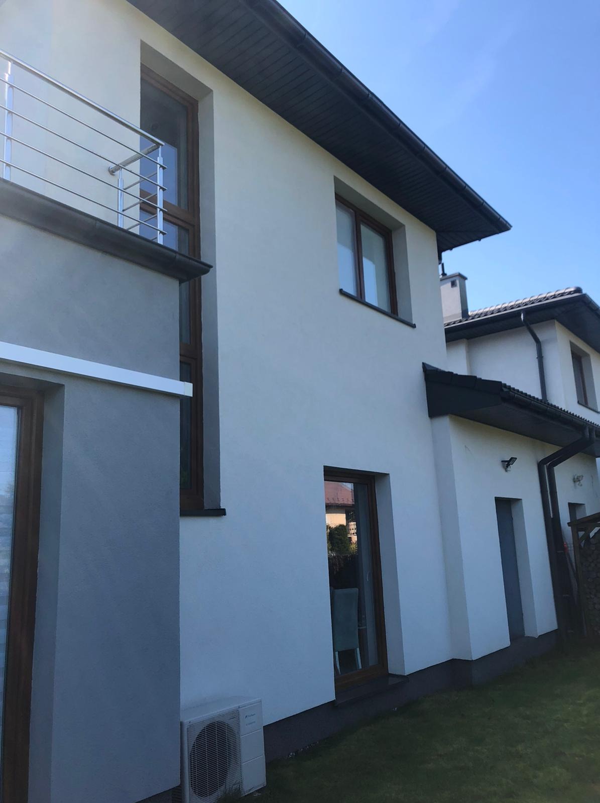 Nowoczesny dom jednorodzinny z jasną elewacją i ciemną stolarką okienną, widoczny klimatyzator na ścianie, balkon z metalową balustradą.