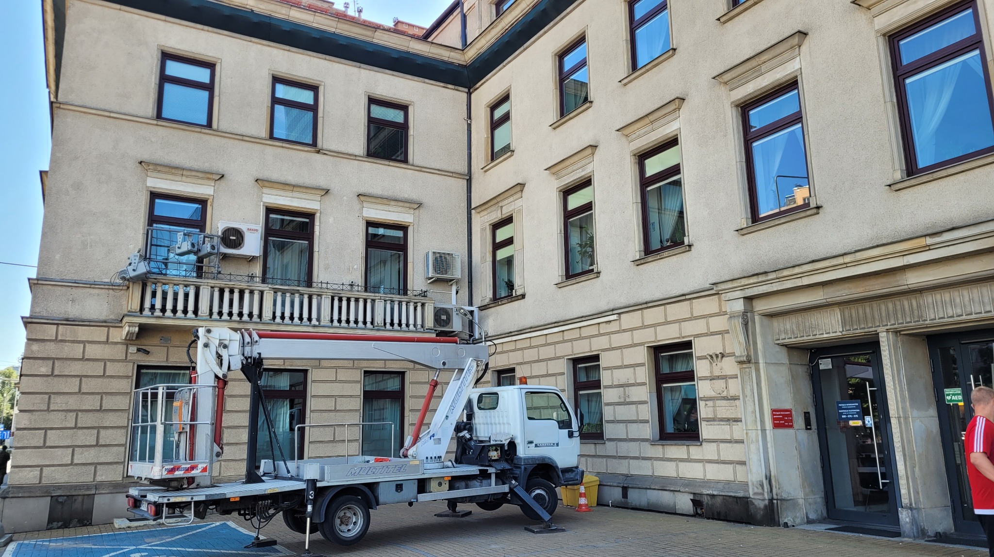 Podnośnik koszowy na tle jasnej, dwupiętrowej elewacji budynku z licznymi oknami i balkonem, przygotowany do prac na wysokościach.