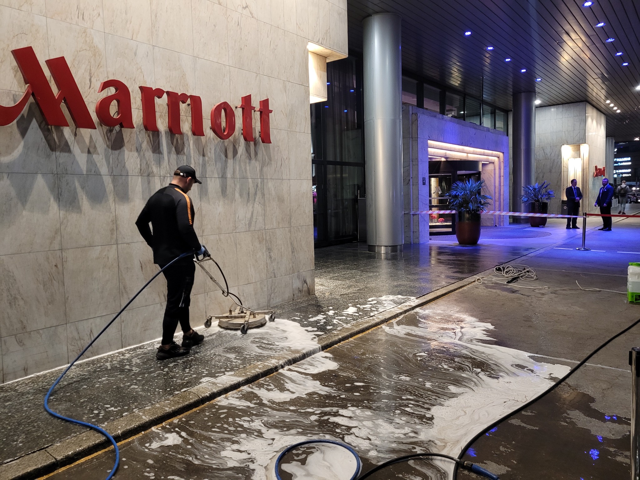 Pracownik w czarnym stroju czyści chodnik przed budynkiem z czerwonym logo za pomocą myjki ciśnieniowej, tworząc pianę na powierzchni.