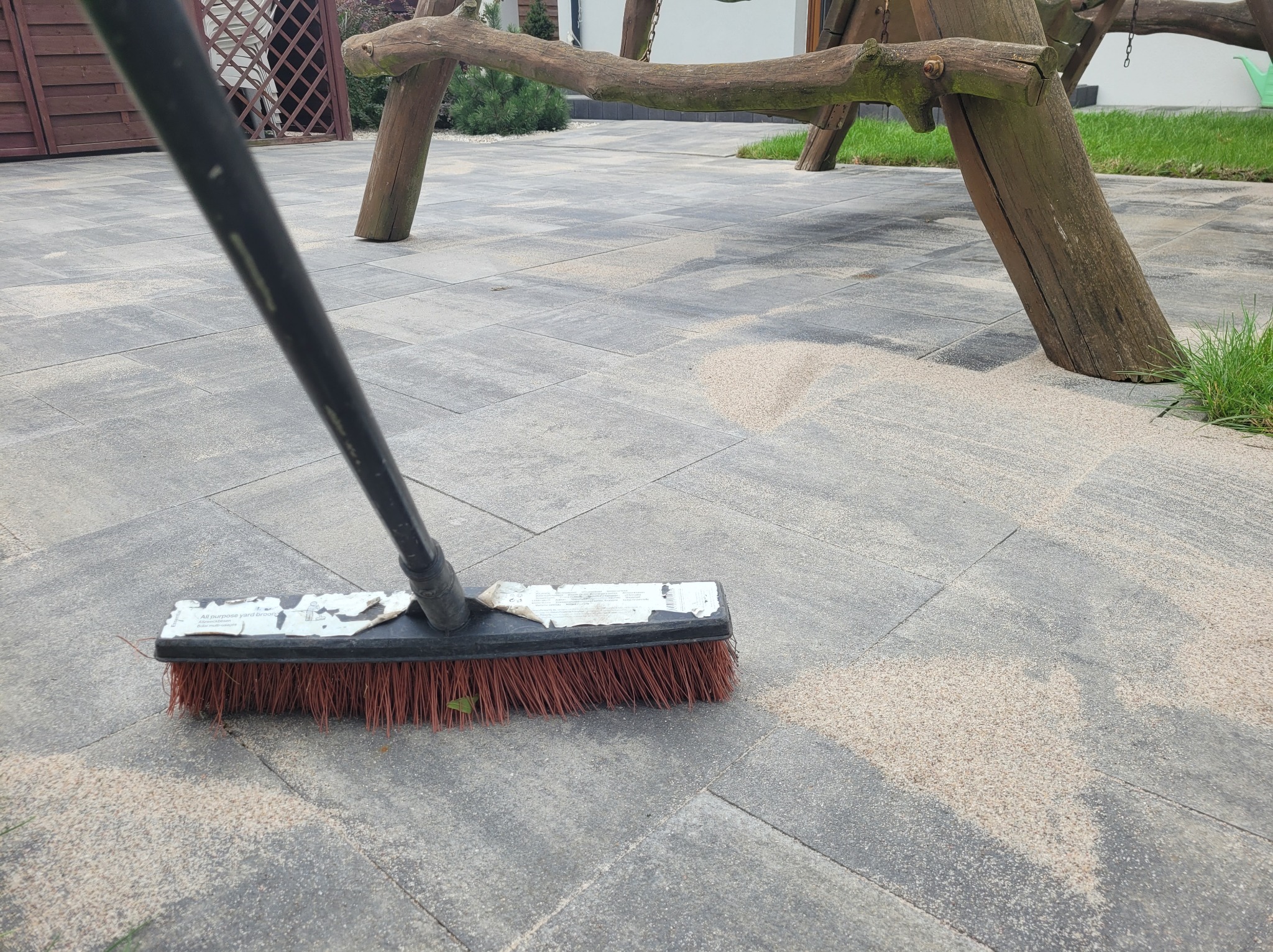 Szczotka zamiata piasek z szarej kostki brukowej na podwórku z drewnianą huśtawką w tle.