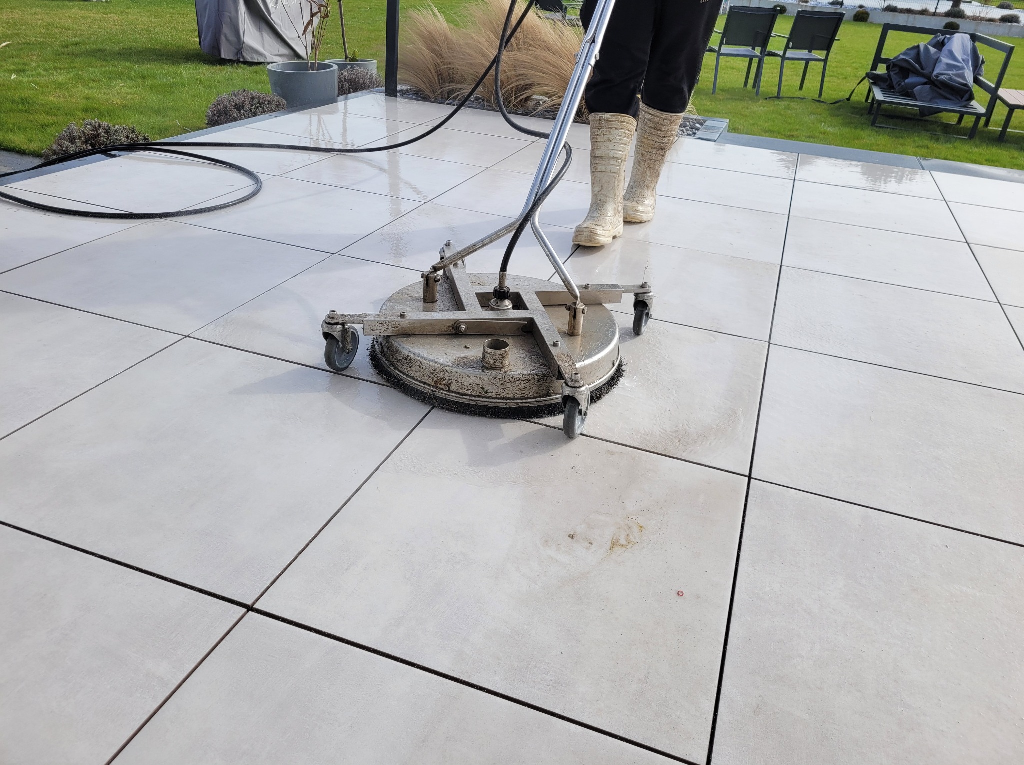 Pracownik w beżowych gumowcach czyści jasne płytki tarasowe za pomocą rotacyjnej myjki ciśnieniowej na tle zadbanego trawnika i mebli ogrodowych.