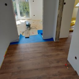 Grzegorz Bitner - Montaż paneli podłogowych w trakcie remontu, widoczna piła elektryczna, niebieska folia ochronna, oraz paczka nożyków do tapet na brązowej podłodze.