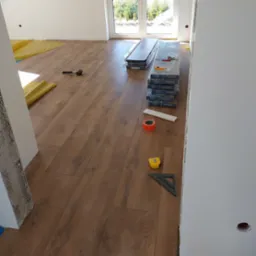 Układanie paneli podłogowych w jasnym pomieszczeniu z widokiem na zewnątrz, widoczne narzędzia: miarka, kątownik, młotek gumowy oraz paczki paneli marki Megaloc.