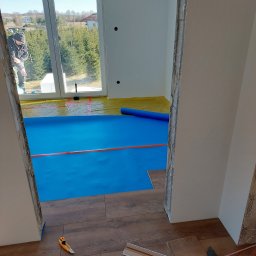 Grzegorz Bitner - Montaż paneli podłogowych w pokoju z widokiem na zewnątrz, zabezpieczona folia pod spodem, widoczne narzędzia i listwy przypodłogowe.