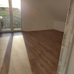 Grzegorz Bitner - Pokój na poddaszu po remoncie: panele podłogowe w odcieniach brązu, białe ściany i widok na zewnątrz przez okno z widocznymi promieniami słonecznymi. Na pierwszym planie listwa przypodłogowa...