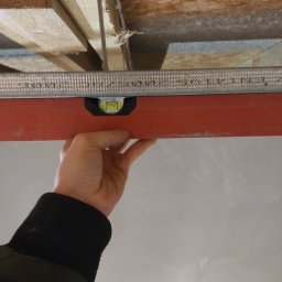 Grzegorz Bitner - Ręka przytrzymuje poziomicę na metalowym profilu pod drewnianym stropem, widoczny fragment sufitu w trakcie prac wykończeniowych.