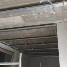 Montaż metalowego rusztu pod płytę karton gips na surowym betonowym suficie, widoczne profile i uchwyty montażowe, rusztowanie malarskie w pomieszczeniu.