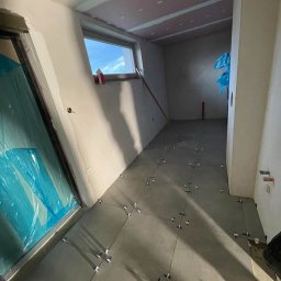 Grzegorz Bitner - Wnętrze w trakcie remontu: szare płytki podłogowe z krzyżykami dystansowymi, ściany przygotowane do malowania, okno z widokiem na niebo, zabezpieczone folią drzwi.