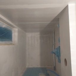 Grzegorz Bitner - Przygotowane do malowania pomieszczenie z zabezpieczonym oknem i podłogą folią malarską, ściany po szpachlowaniu, widoczny fragment rury również zabezpieczony folią.