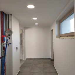 Grzegorz Bitner - Korytarz w trakcie remontu z widocznymi instalacjami, świeżo pomalowanymi ścianami i zamontowanymi lampami sufitowymi, okno z widocznymi śladami po pracach wykończeniowych.
